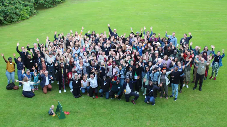 A large group of people stand in a green space and look up waving at a camera. 