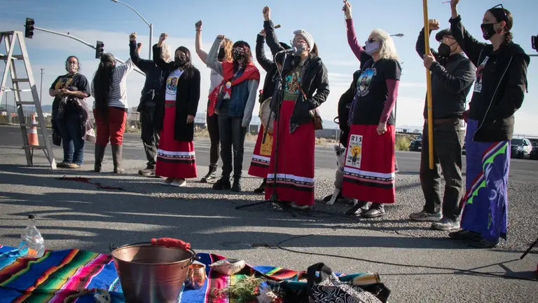 A group of Indigenous Women wearing traditional dress protesting outside, holding signs and raising their fists in the air