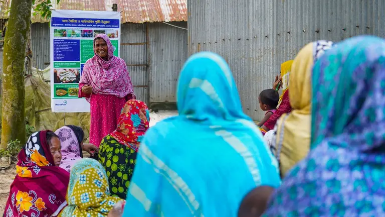 Group of women gathered for a regular meeting on aquaculture. One woman is standing and speaking with a poster behind her, while a groups is sitting and listening.