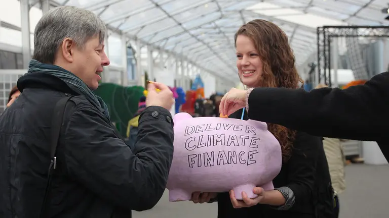 A woman holds a pink piggy bank in front of her, with the words 'Deliver climate finance' written on the front.