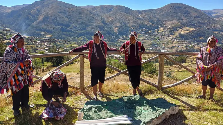 A group of Indigenous farmers, wearing brightly coloured clothes, smile as they lean on a wooden rail and gather around a large rock, in front of a scenic landscape of mountains under a blue sky.