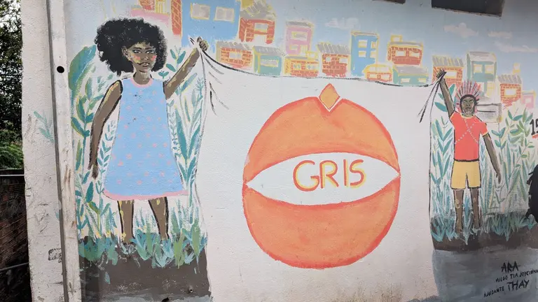 A mural on the wall of two children holding a big banner that reads 'Gris'. One of them is wearing a bright blue dress and the other a red shirt and yellow shorts. They faces are covered in paints.