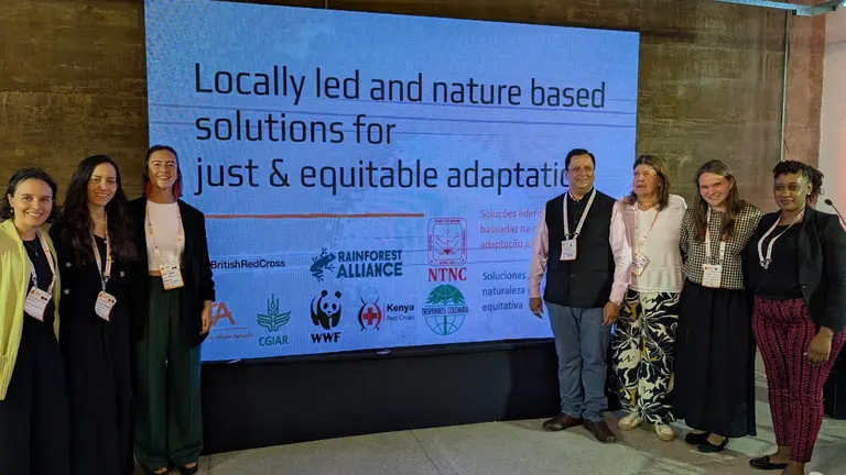 People standing together for a group photos. Behind them, a big screen is projecting a PowerPoint presentation.