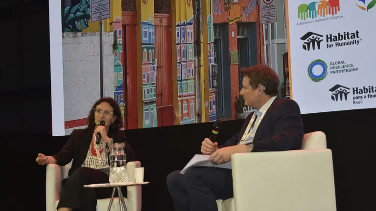 Two people sitting down on a stage, holding microphones and discussing.
