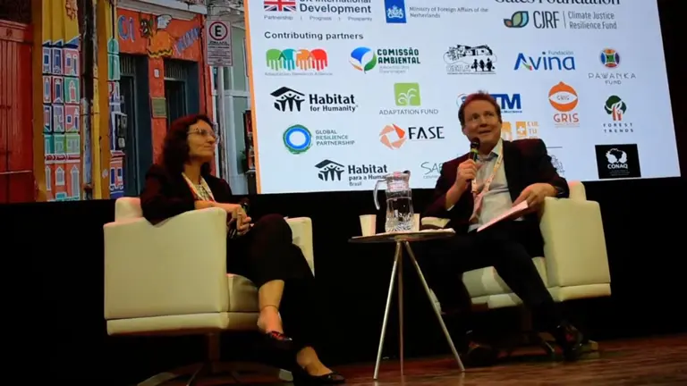 Two people sitting down on a stage, holding microphones and discussing.