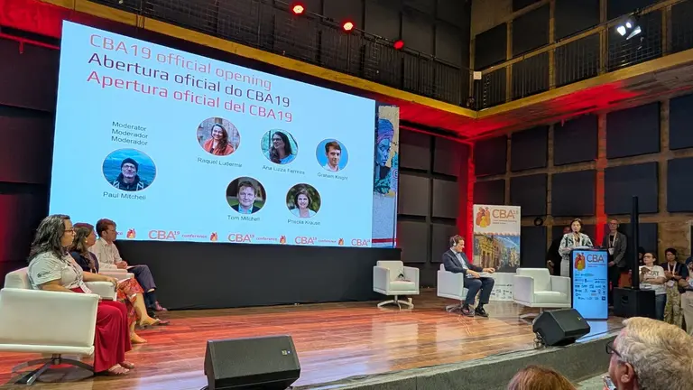 Image of stage at CBA19 conference centre with four people sitting on white chairs and one person speaking. There is a big screen behind projecting a PowerPoint presentation.