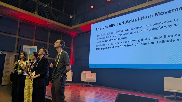 Four speakers are standing on a stage, looking at the audience. Behind them is a projection of a PowerPoint presentation.