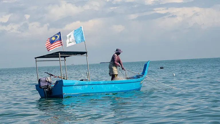 A small-scale fisherwoman stands in a small blue boat with a net flowing into the water.