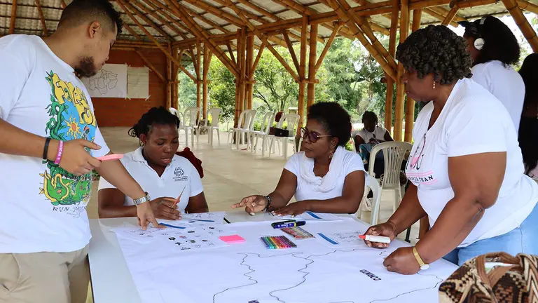 People sit and stand around a table and add comments to a white piece of table on the table