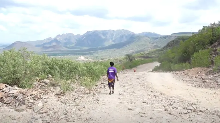 A woman walks alongside a dirt-track road with mountains in the background.