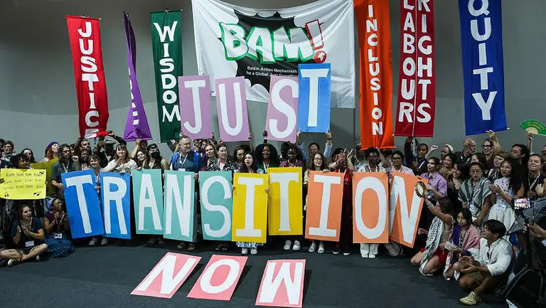 A large group of people carrying signs make the words 'Just transition now' by holding up letters on boards. 