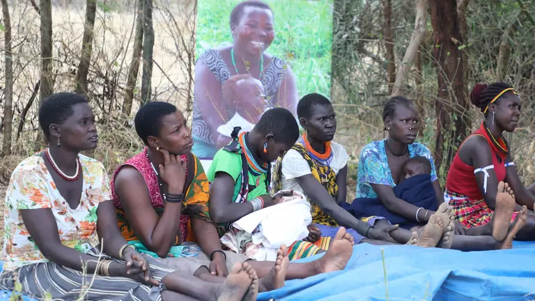 Women sit on blankets on the floor as part of a discussion, with trees and a display board behind.