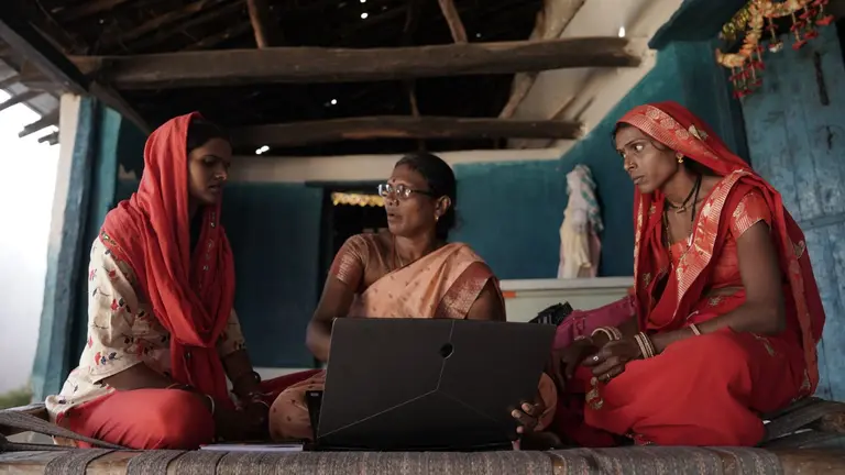 Three women wearing tradition sarees sitting down and discussing. One of them is holding a laptop and explaining something to the other two women.