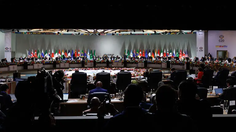 In the foreground, the backs of people watching a long line of people seated at a brightly lit panel in the distance.