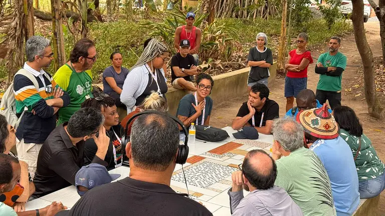A group of standing and seated people gather outside around a table, on which is laid out paper featuring maps and other items, with trees in the background.