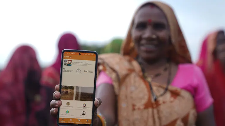 A woman farmer holds a mobile phone showing the CRISP-M tool towards the camera.