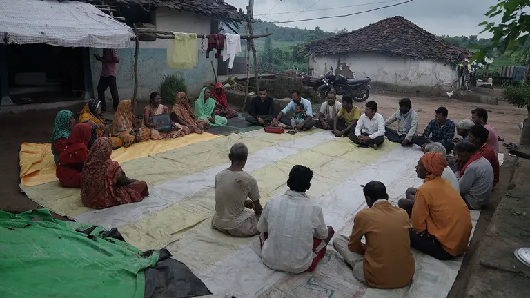 A group of people seated in a circle on blankets outside have a discussion, with one woman showing a laptop screen to everyone else.