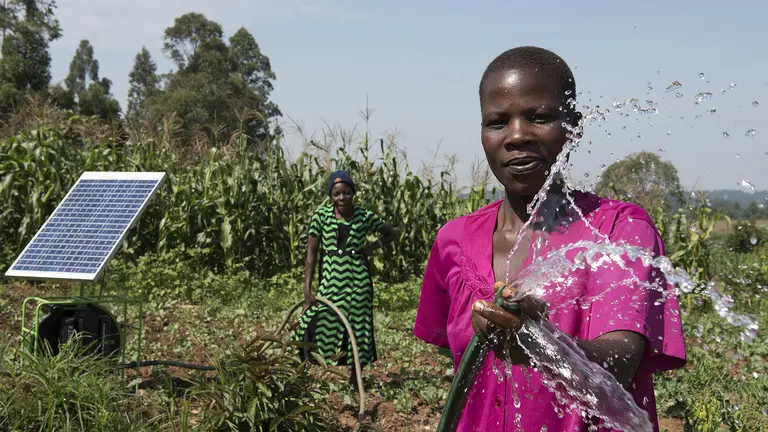 A woman farmer sprays water out of a hosepipe with a solar panel in the field behind her. 