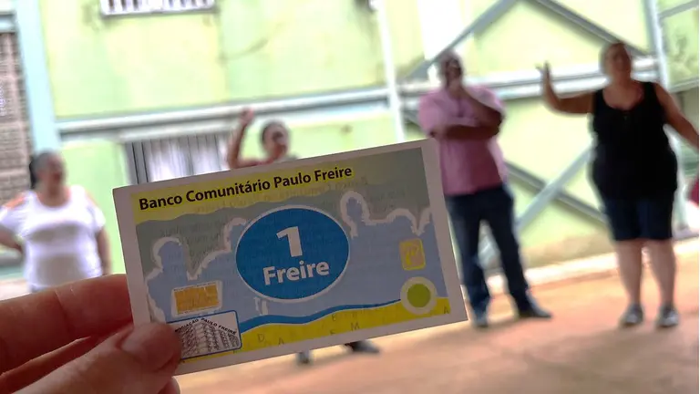 A hand holds a paper voucher, saying "1 Freire" close to the camera while people are blurred in the background. 