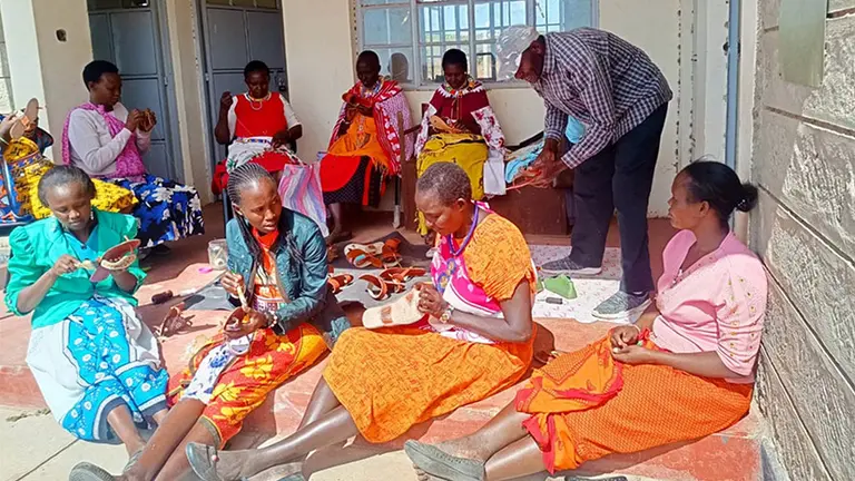 Women sit on the floor and in chairs as they carefully stitch sandals.