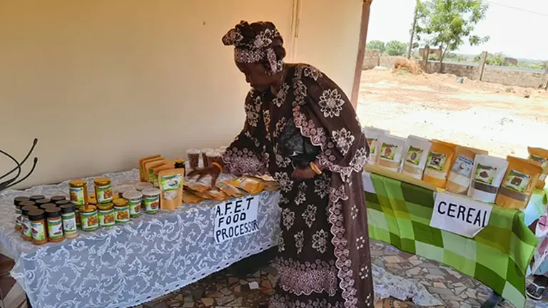 A woman bends over a table stocked with small pots/products to sell. A table behind is labelled 'cereal'.