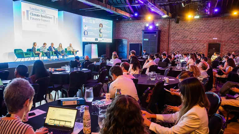 Hundreds of people, seated at small tables, listen to people seated in a panel-style format on a main stage at a conference.
