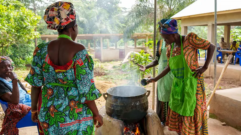 Women in brightly coloured clothes gather around a large smoking pot outside.