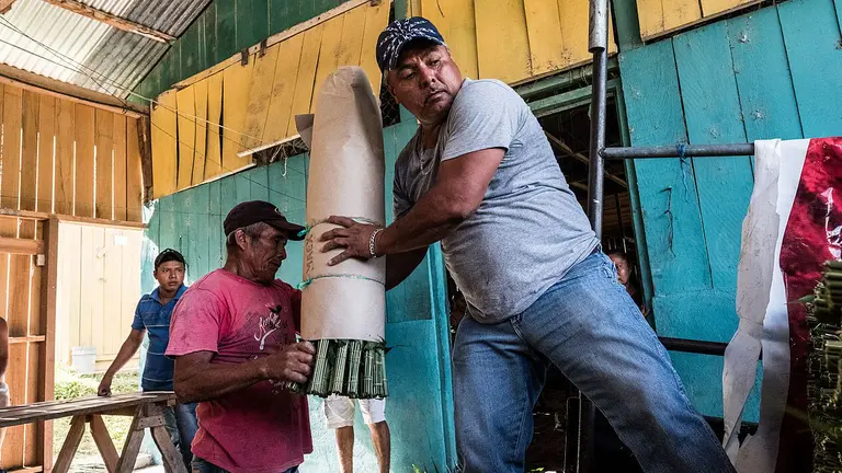 Two men lift and together carry a bundled package in a small factory-type room.