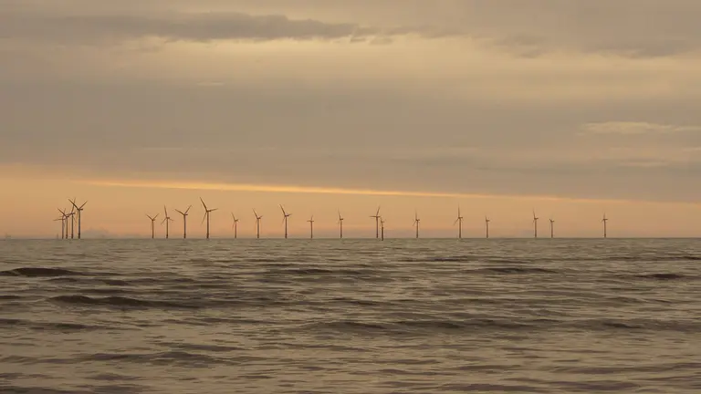 A dramatic sunset of reds and yellows frames a dozen wind turbines in the distance in the water.