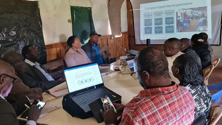 A group of people sit around a table looking at computers screens and mobile phones while receiving a presentation, that is being shown on a big screen.