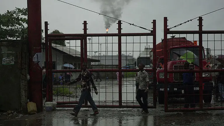 People walk past imposing dark gates, behind which can be seen an oil refinery. 