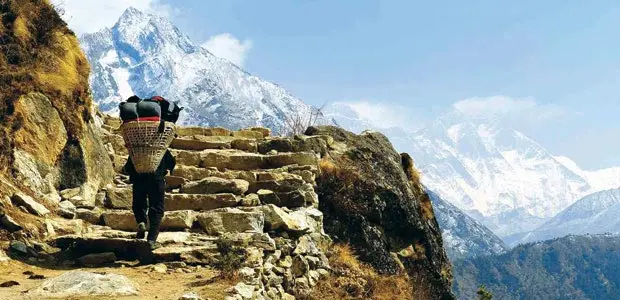 A Nepalese man carrying a large sack on his back walks up some steps with the Himalayas soaring behind. 