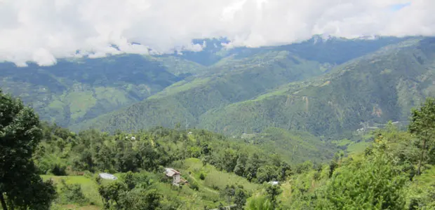 Forested hills in Nepal, with a house in the foreground and forested hills, and then clouds behind.