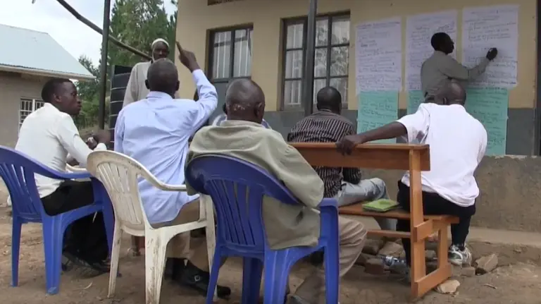 People sitting on outdoor chairs gather around and contribute views as one person writes up their points on paper pinned to the wall