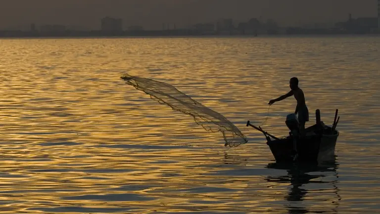A fisher casting a net from a small boat surrounded by a large expanse of water 