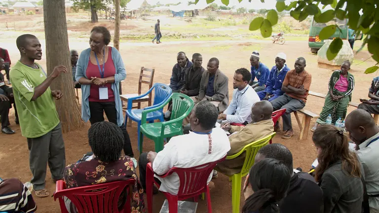 A man stands as he speaks to a seated group of people surrounding him