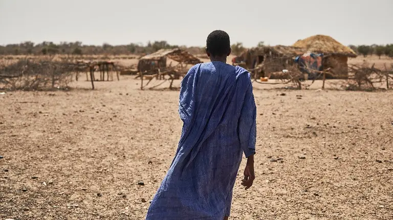 Man, wearing a blue tunic, stands with his back to the camera and looks at huts in the background.