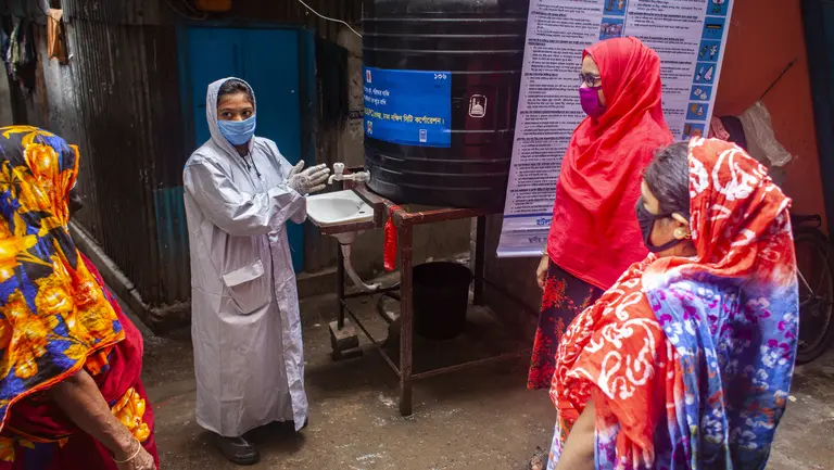 A woman covered in protective plastic and wearing a mask demonstrates handwashing to other women outside.