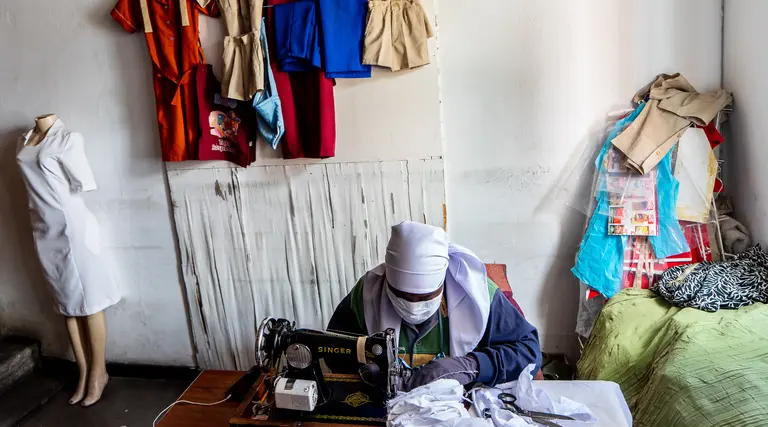 A seated woman, wearing a face mask, huddles over a sewing machine making face masks.
