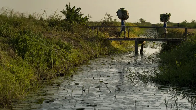 Two people carrying bags on their head walk through a bridge over a river
