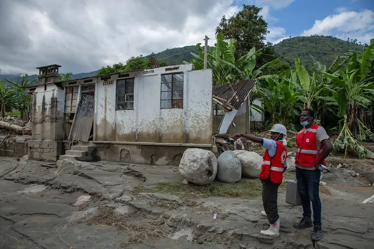 Two men with vests from the Red Cross in front of a destroyed building