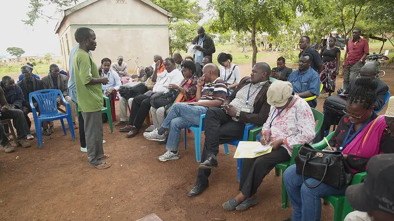 A man stands in front of a group of seated people outside answering questions