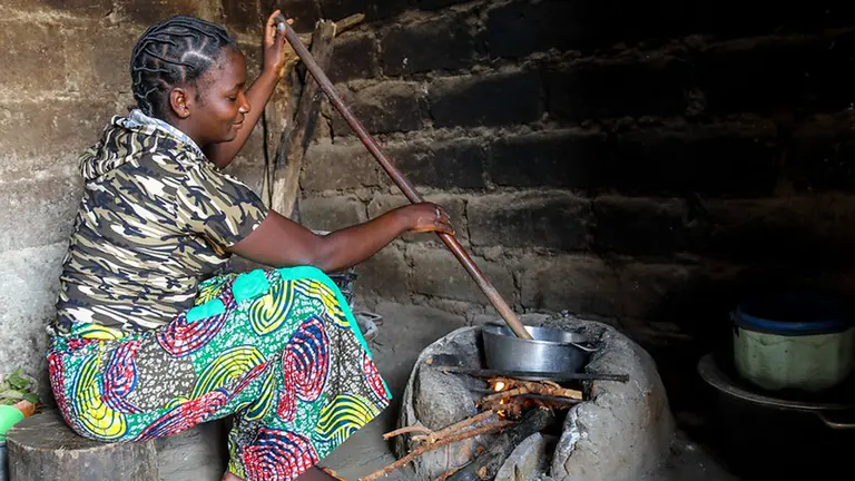 Woman stirs a long stick in a pan on a fireplace made of stone