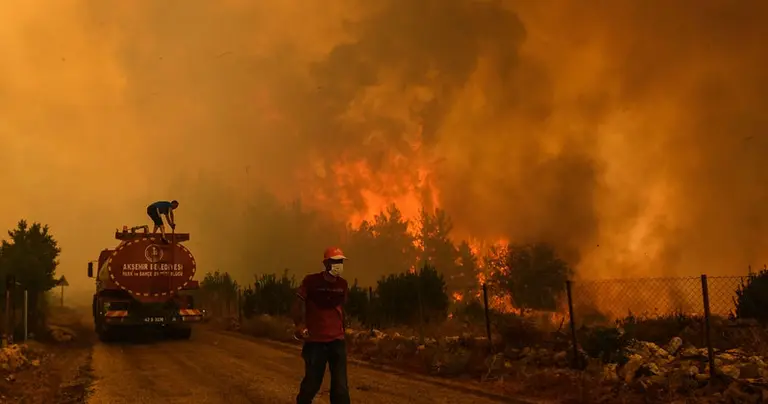 A wildfire burns a forest. A man climbs a fire engine and another one, wearing a mask, walk on the road. 