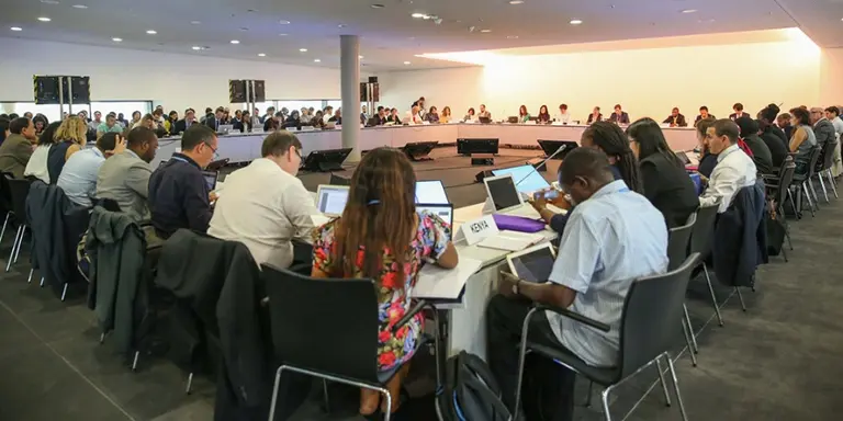 A large number of people, with backs to the camera, are seated around a table. On the tables there are cards indicating which countries they represent
