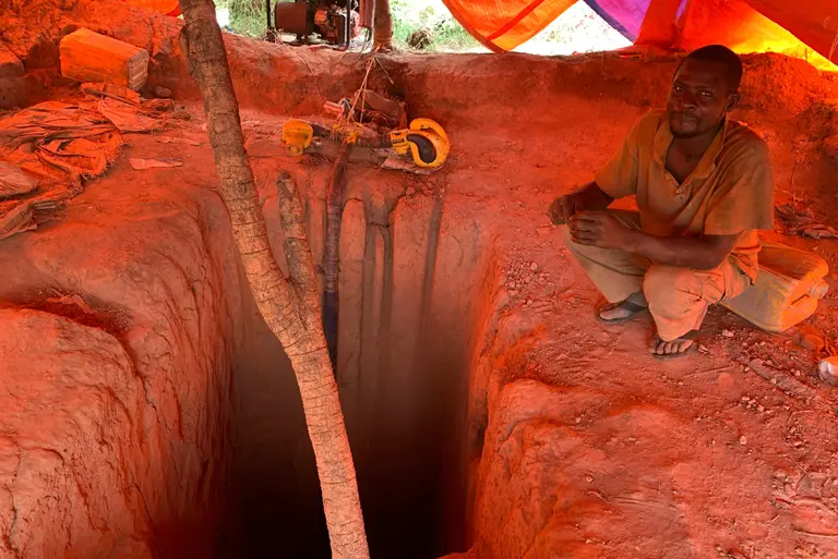 A man crouches on his haunches under canvas next to a large hole in the ground