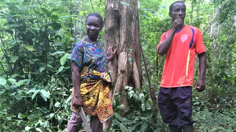 A man and a woman holding a machete stand in front of a tree in a forest