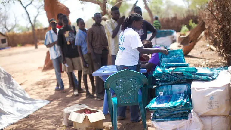 Men queue in front of a table. A woman hands them bags.