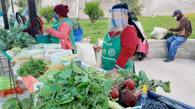 A woman wearing a face mask and a visor stands behind a table of vegetables selling her produce.
