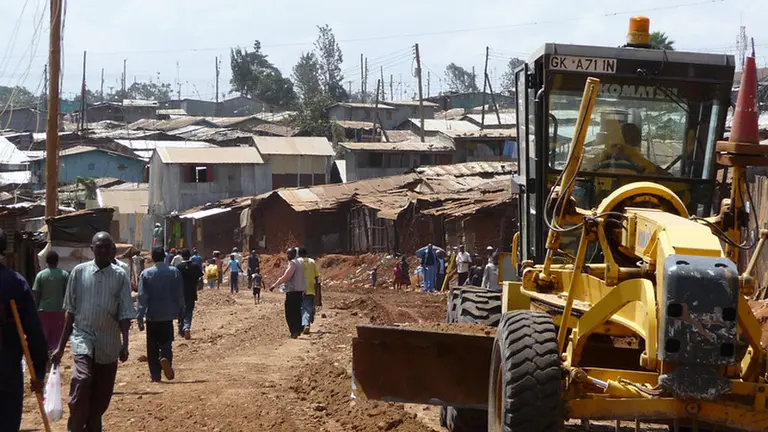 Large yellow digger vehicle parked in front of slum dwellings. 